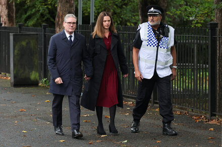 Keir Starmer, leader of the Labour Party, stands outside a synagogue in Manchester, U.K., with serious expression, surrounded by security. The synagogue's exterior is visible behind him. The image is from a news report.
