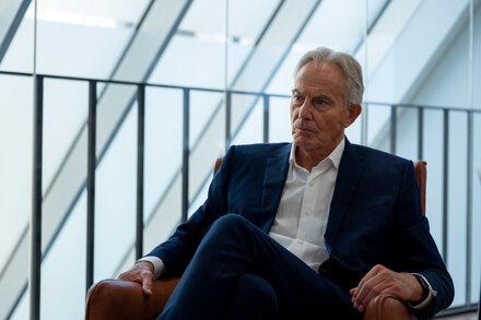 El ex primer ministro británico Tony Blair camina con un grupo de personas en un entorno que parece ser de Oriente Medio, posiblemente en Gaza, con edificios dañados o en construcción. Viste una camisa clara y pantalones oscuros. La imagen tiene un tono serio y contemplativo.
