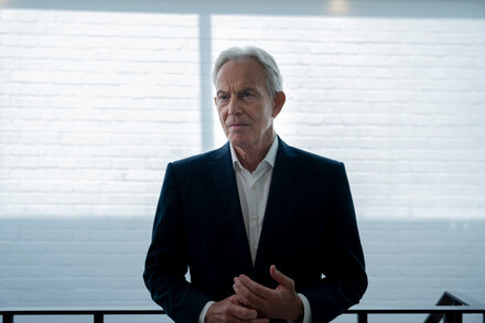 Tony Blair, former UK Prime Minister, stands in front of a blue background, looking serious. He wears a dark suit and light blue tie. The lighting emphasizes his face.