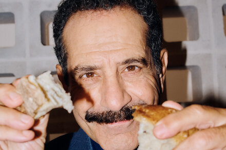 Tony Shalhoub, with a warm smile, holds up a piece of artisanal bread against a rustic backdrop, showcasing his enjoyment of culinary travel.