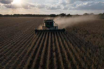 President Trump speaking at a rally, with farmers in attendance and agricultural fields visible in the background, symbolizing his administration's support for the farming community amidst trade tensions.