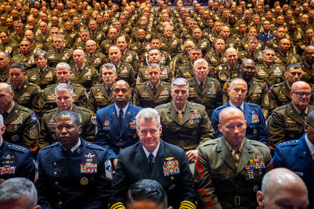 Donald Trump speaks at a podium with Pete Hegseth standing nearby, addressing a group of uniformed senior U.S. military officers seated in an auditorium in Virginia.