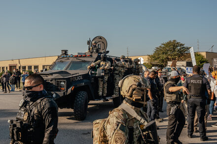 Donald Trump speaks at a podium with a serious expression, gesturing with his hand, as military personnel stand in the background, symbolizing his threat to deploy the National Guard under the Insurrection Act.