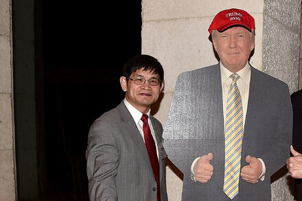 Donald Trump speaks at a rally while the Department of Justice building is visible in the background, symbolizing the ongoing legal pursuit of his adversaries and a political ally's trial.