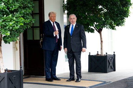 Donald Trump and Benjamin Netanyahu speak at a podium with microphones in front of a blue background with flags.