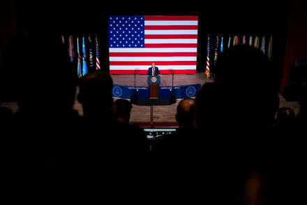 Donald Trump speaking at a podium with a military backdrop, gesturing with his hands. The image captures him mid-speech, appearing to make a point.