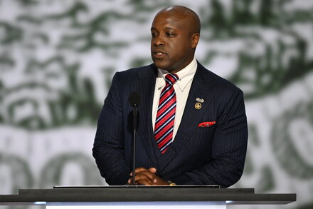 Wesley Hunt, an African American man in a suit, stands at a podium with the Texas flag in the background, addressing a crowd.