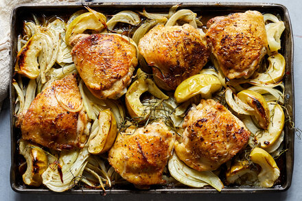 A roasted chicken dish with cooked apple slices and herbs on a white plate, garnished with fresh herbs and a lemon wedge.